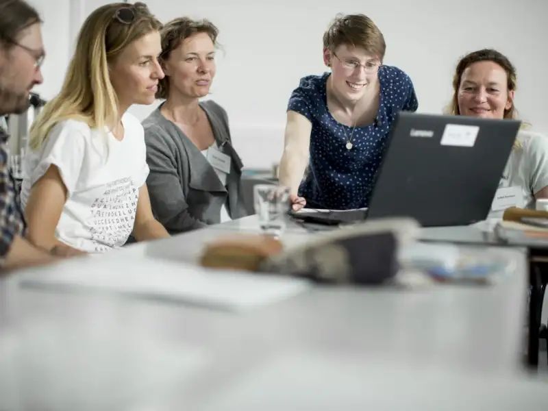 Mehrere Personen sitzen an Schultischen und gucken gemeinsam auf einen Laptop.