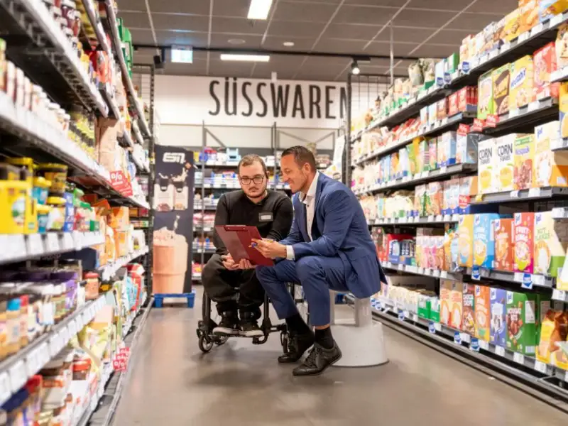 REWE Supermartkbetreiber David Hegemann und sein Auszubildenden Achilleas Tsantekidis, der im Rollstuhl sitzt, vor den warenregalen im Supermarkt.