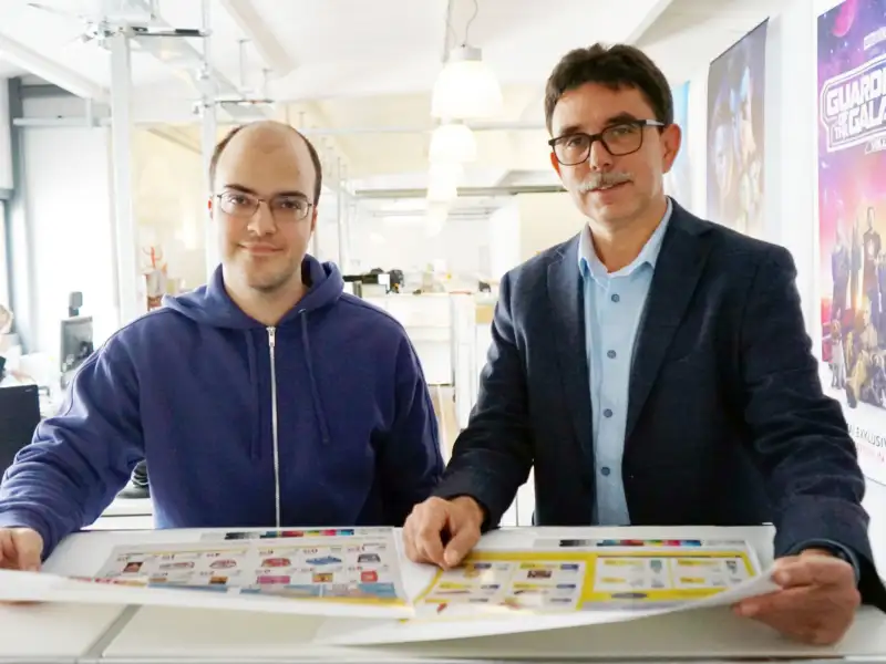 Dennis Niecznik (links) und sein Chef Guido Richter stehen nebeneinander an einem Arbeitstisch der Werbeagentur.