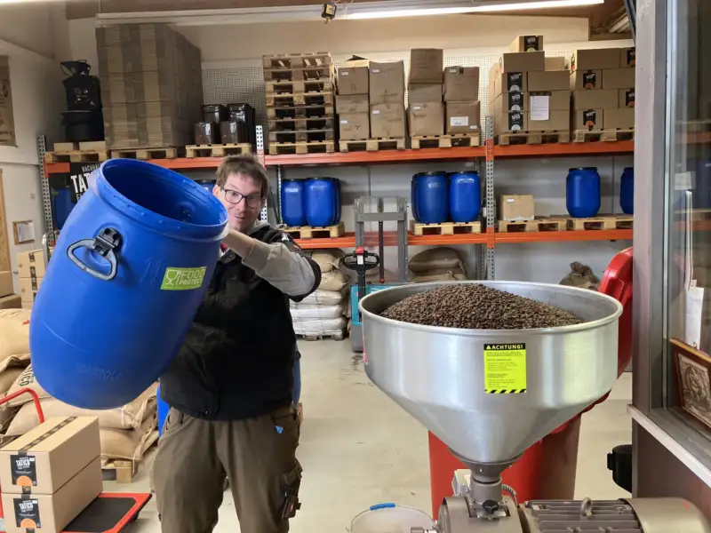 Mitarbeiter Marvin Tutsch schüttet Kaffebohnen aus einem Fass in eine große Kaffeemühle.