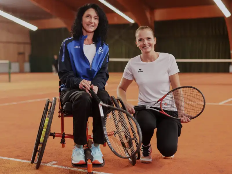 Zwei Tennis-Spielerinnen in einer Tennis-Halle, eine ist Fußgängerin, die andere Rollstuhlfahrerin.