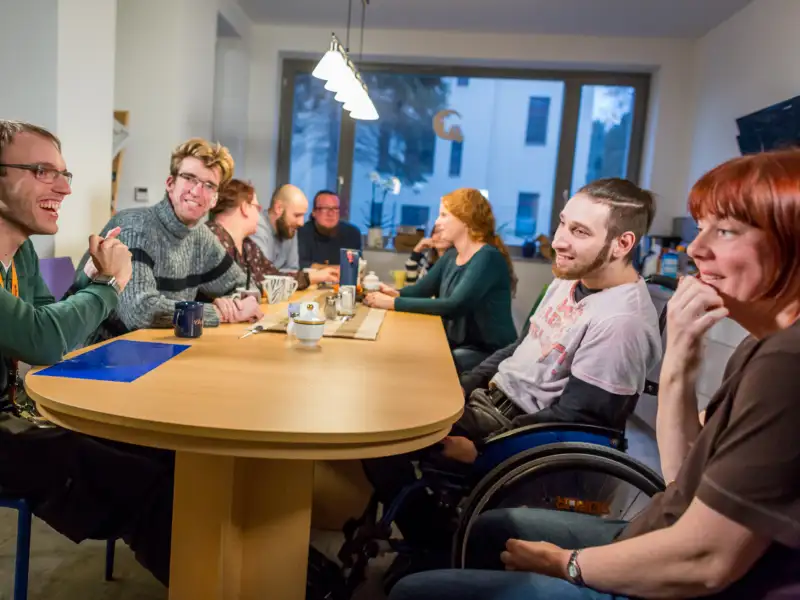 An einem Esstisch sitzen viele verschiedene Menschen mit und ohne Behinderung.