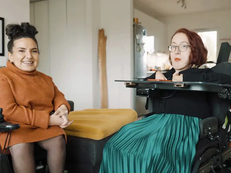 Zwei junge Frauen im E-Rolli sitzen nebeneinander in einer Wohnung