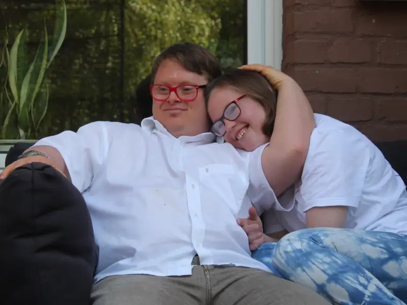 Ein junges Pärchen - beide mit Downsyndrom - sitzen auf dem Sofa. Er hat seinen Arm um sie gelegt und sie kuschelt sich an ihn an.