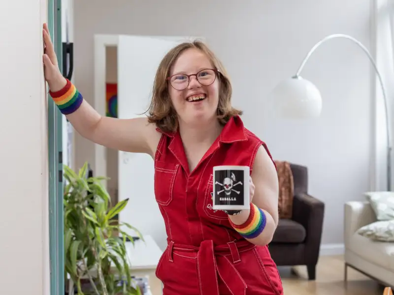 Natalie Dedreux steht in einem roten Jumpsuite in einem Wohnzimmer und lacht in die Kamera. Sie hält sich mit der rechten Hand an der Wand fest.