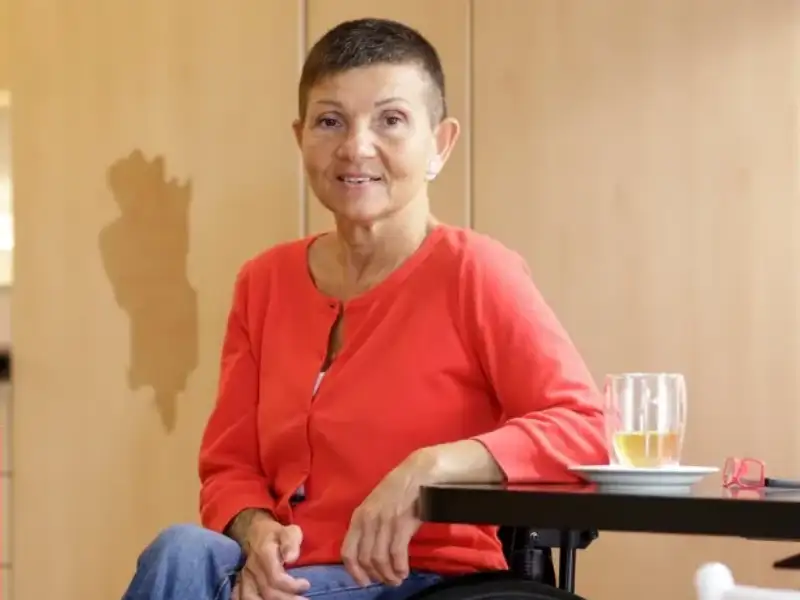Silvana sitzt im Rollstuhl an einem Tisch. Sie trägt ein rotes Oberteil, eine blaue Jeans und rote Turnschuhe. Neben ihr steht ein Glas mit einem Getränk.