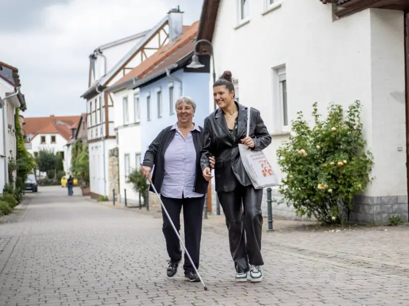 Zwei Frauen laufen eine Straße entlang, eine Frau nutzt einen Blinden-Langstock.