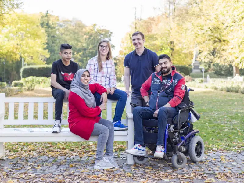 Fünf Menschen sitzen und stehen um eine Bank in einem Garten, ein Mann sitzt im Rollstuhl.