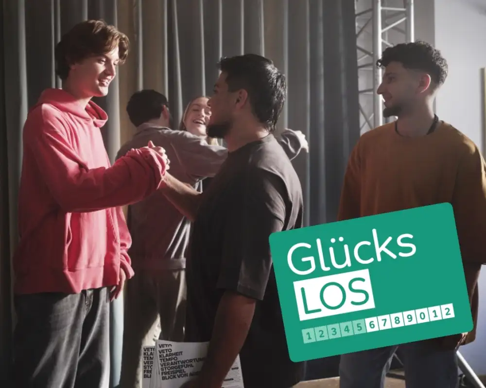 Eine Gruppe Jugendlicher steht auf einer Theaterbühne. Im Vordergrund schlagen zwei von ihnen freundschaftlich miteinander ein. In der rechten unteren Ecke des Bildes ist das grüne Glücks-Los der Aktion Mensch zu sehen.