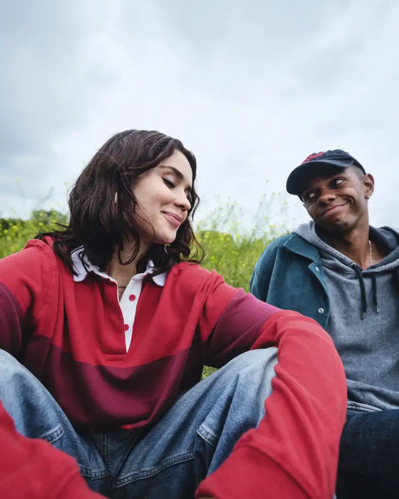 Zwei Jugendliche sitzen auf einer Wiese. Links ist ein Mädchen mit rotem Langarmshirt und Jeans. Rechts ist ein Junge mit grauer Kapuzenjacke, blauer Jacke und Baseballkappe. Sie schauen sich freundlich an.