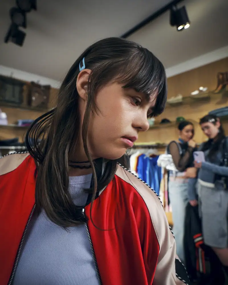 Eine junge Frau mit Downsyndrom steht in einem Bekleidungsgeschäft. Sie schaut unsicher über ihre linke Schulter. Im Hintergrund sind zwei weitere junge Frauen erkennbar, die scheinbar miteinander tuscheln.