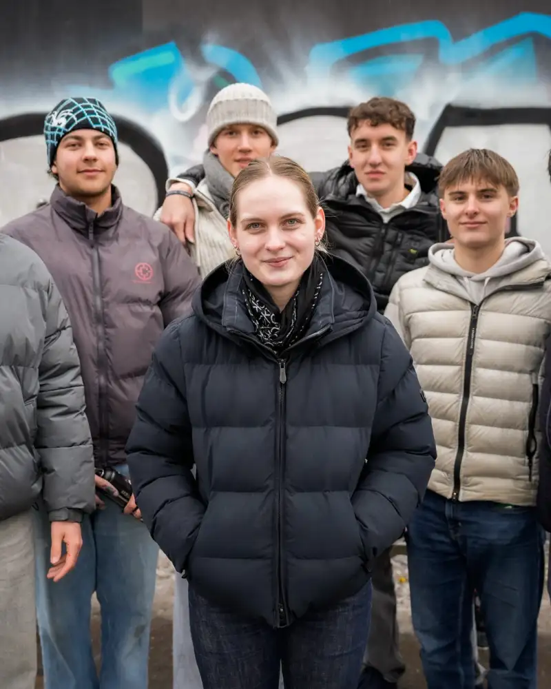 Vor einer mit bunten Graffitis besprühten Wand steht eine Gruppe Jugendlicher. Sie schauen freundlich zur Kamera.