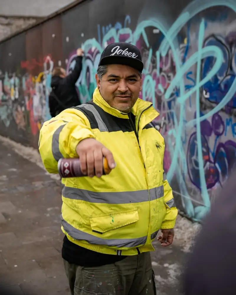 Vor einer mit bunten Graffitis besprühten Wand steht ein Mann in einer grellgelben Jacke. Er hält eine Spraydose vor sich in die Luft und schaut selbstsicher in die Kamera. Im Hintergrund sieht man einen Jugendlichen. Er ist dabei, ein Bild an eine Mauer zu sprayen.