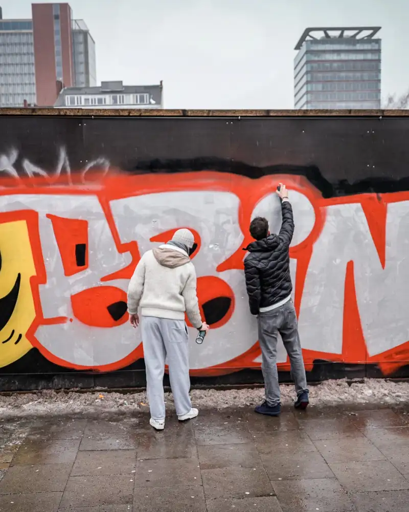 Vor einer mit bunten Graffitis besprühten Wand stehen einige Jugendliche. Sie sind dabei, ein neues Bild an die Wand zu malen. Der Schriftzug Mobbing ist zu lesen.