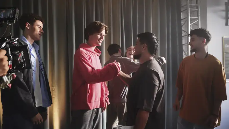 Eine Gruppe Jugendlicher steht auf einer Theaterbühne. Im Vordergrund schlagen zwei von ihnen freundschaftlich miteinander ein.