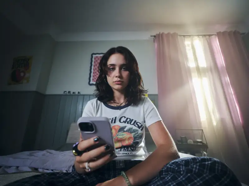 Eine Jugendliche sitzt auf einem Bett in einem düsteren Zimmer. In der Hand hält sie ein Smartphone. Sie schaut traurig am Handy vorbei in die Kamera.