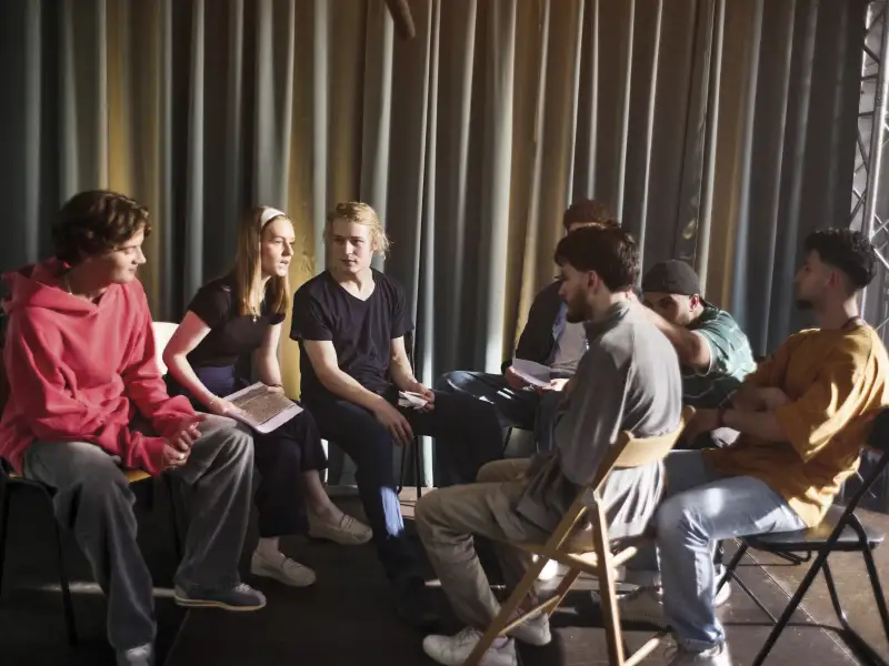 Eine Gruppe Jugendlicher sitzt auf einer Theaterbühne. Zwei von ihnen unterhalten sich miteinander, die anderen schauen interessiert zu.