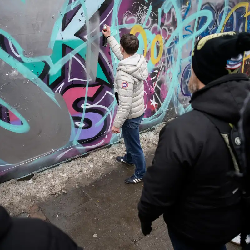 Vor einer mit bunten Graffitis besprühten Wand steht eine Gruppe Jugendlicher. Einer vomn ihnen sprüht gerade mit einer Dose Farbe an die Wand, die anderen schauen zu.