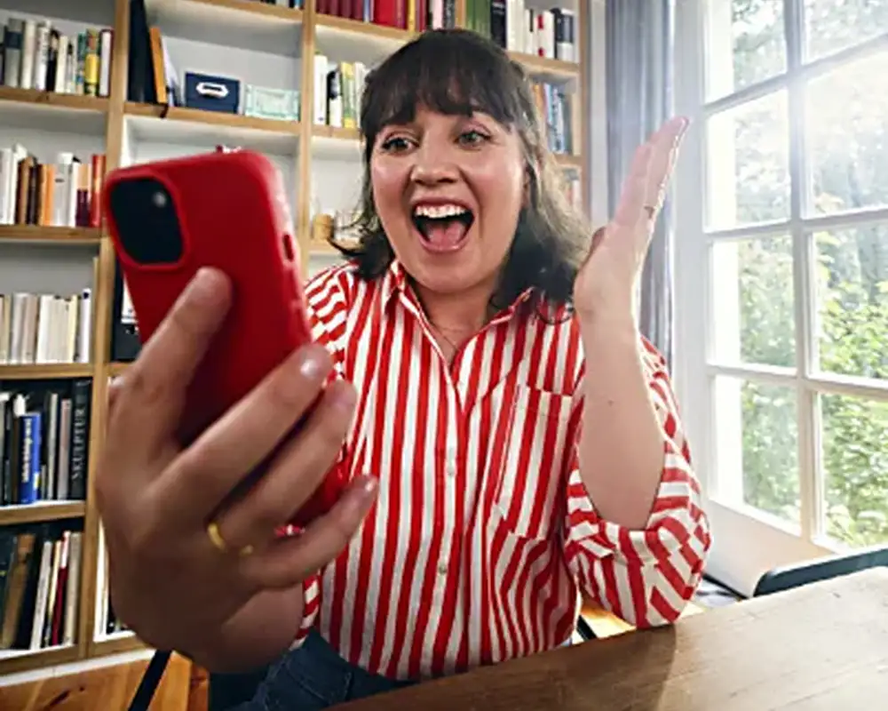 Eine Frau in einem rot-weiß gestreiften Hemd sitzt an einem Tisch in einem hellen Raum und hält ein rotes Smartphone in der Hand. Sie freut sich offensichtlich über etwas und gestikuliert mit ihrer freien Hand, um ihre Emotionen auszudrücken. Neben ihr auf dem Tisch steht ein Globus.