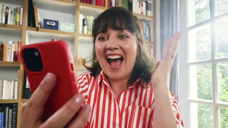 Eine Frau in einem rot-weiß gestreiften Hemd sitzt an einem Tisch in einem hellen Raum und hält ein rotes Smartphone in der Hand. Sie freut sich offensichtlich über etwas und gestikuliert mit ihrer freien Hand, um ihre Emotionen auszudrücken. Neben ihr auf dem Tisch steht ein Globus.