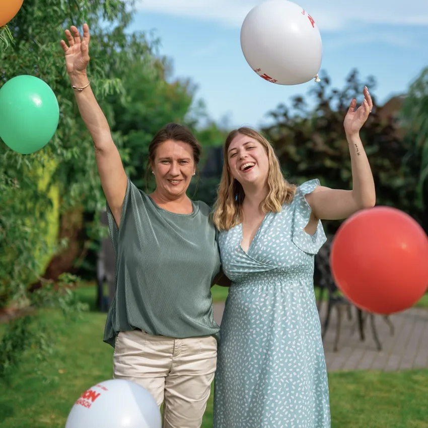 Zwei Frauen in Sommer-Kleidung stehen lachend in einem Garten und werfen Luftballons in die Höhe.