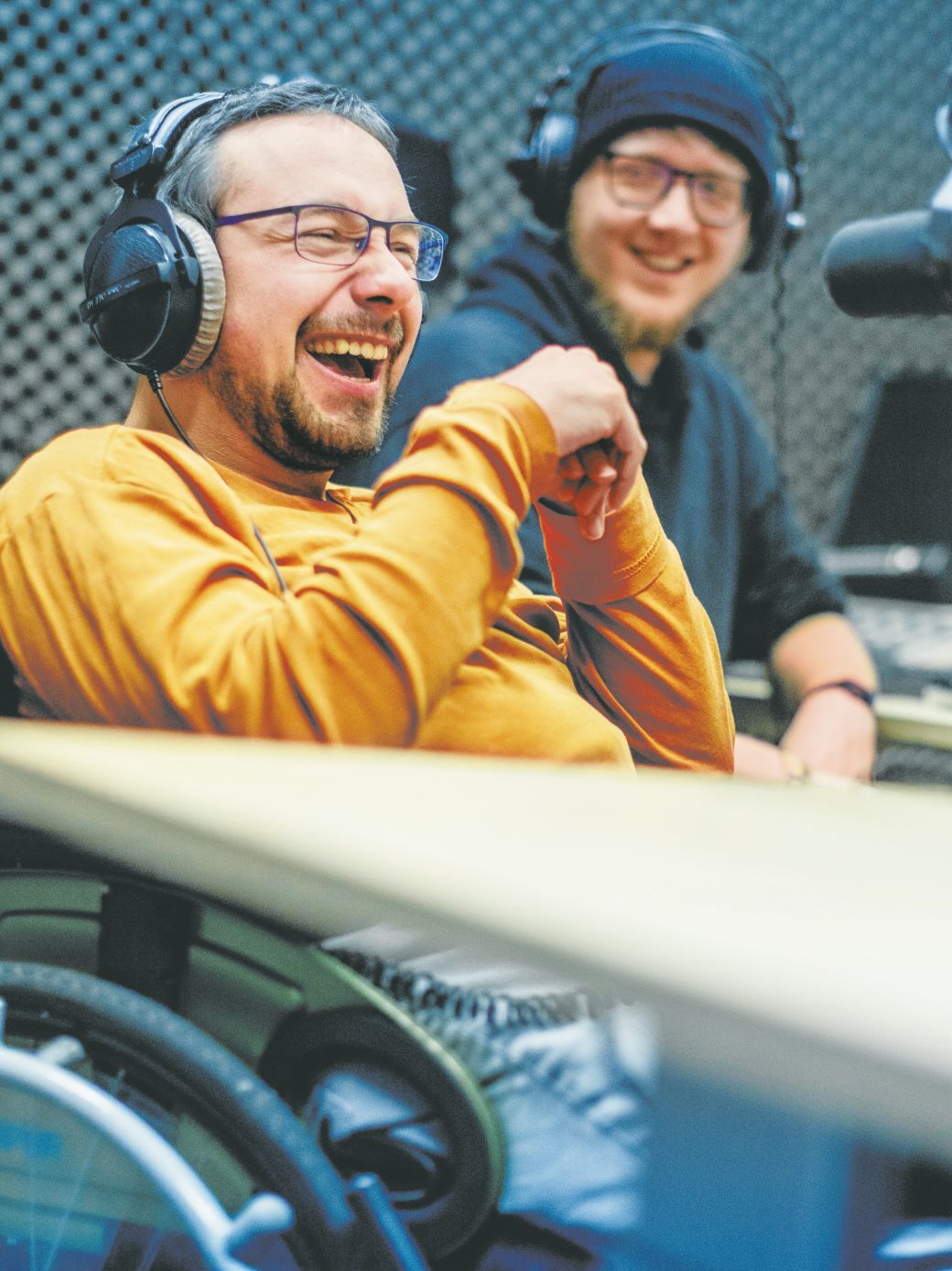 Zwei junge Männer sitzen in einem Radiostudio. Beide tragen Kopfhörer. Einer von beiden nutzt einen Rollstuhl.