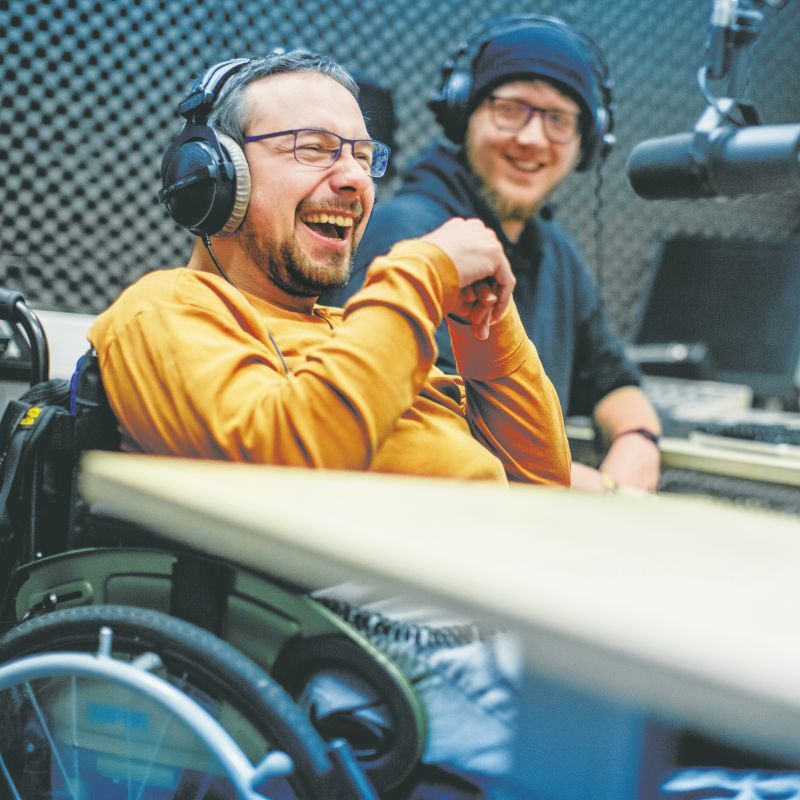 Zwei junge Männer sitzen in einem Radiostudio. Beide tragen Kopfhörer. Einer von beiden nutzt einen Rollstuhl.