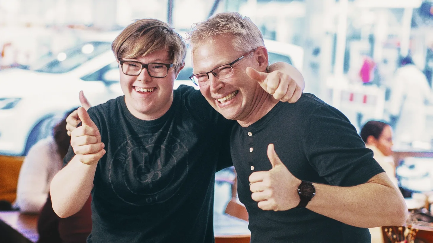 Ein junger Mann mit Down-Syndrom und ein weiterer Mann haben kameradschaftlich die Arme um die Schultern gelegt. Beide lachen und geben das "Daumen hoch"-Zeichen.