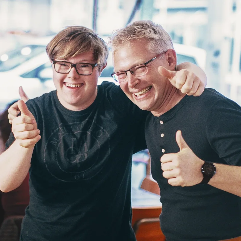 Ein junger Mann mit Down-Syndrom und ein weiterer Mann haben kameradschaftlich die Arme um die Schultern gelegt. Beide lachen und geben das "Daumen hoch"-Zeichen.