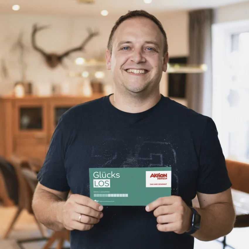 Ein freundlich lächelnder Mann steht in einem gemütlichen Wohnraum und hält ein Glücks-Los der Aktion Mensch in der Hand. Im Hintergrund sind rustikale Holzmöbel und eine stilvolle Inneneinrichtung zu sehen, die eine einladende Atmosphäre schafft.