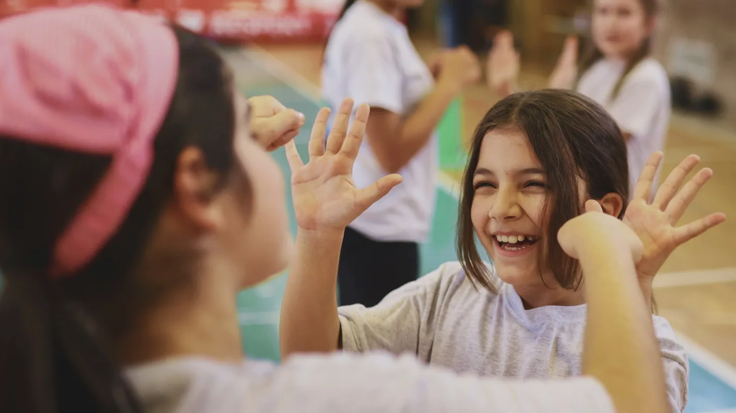 Zwei Mädchen stehen gemeinsam in einer Sporthalle. Sie schlagen miteinander ein und lachen. Beide tragen Freizeitkleidung und scheinen an einer Aktivität oder einem Spiel teilzunehmen. Im Hintergrund sind weitere Kinder zu sehen, die ebenfalls an der Aktivität beteiligt sind.