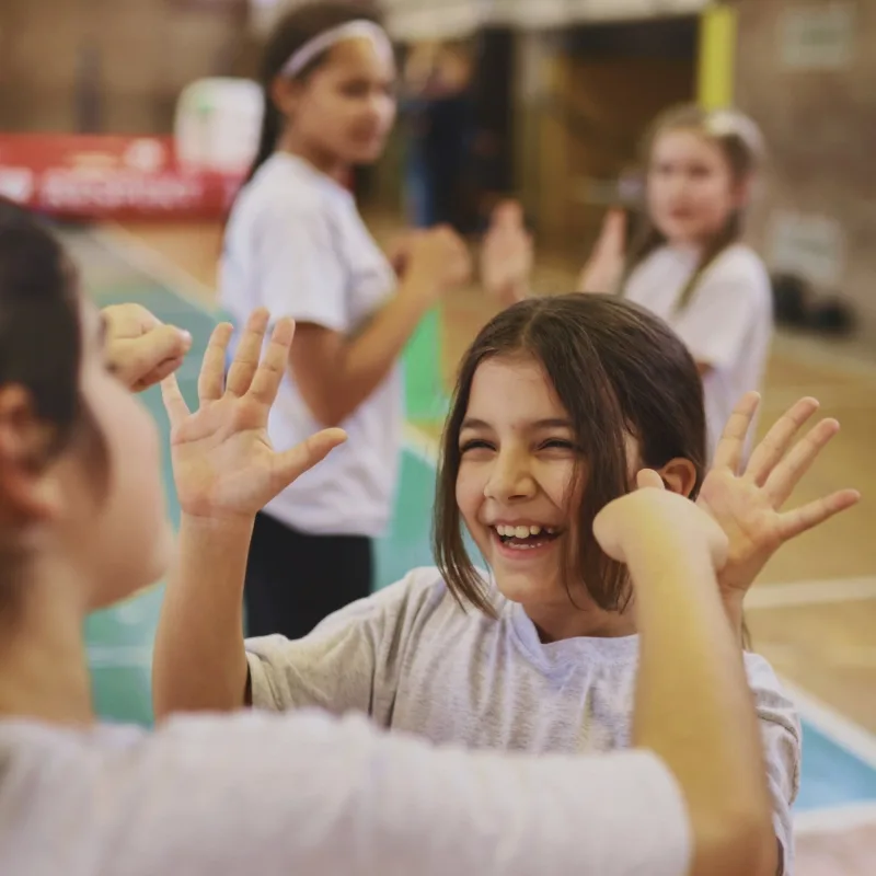 Zwei Mädchen stehen gemeinsam in einer Sporthalle. Sie schlagen miteinander ein und lachen. Beide tragen Freizeitkleidung und scheinen an einer Aktivität oder einem Spiel teilzunehmen. Im Hintergrund sind weitere Kinder zu sehen, die ebenfalls an der Aktivität beteiligt sind.