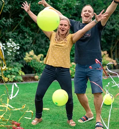 In einem Garten stehen ein Mann, eine Frau und ein Mädchen, alle in sommerlicher Kleidung. Sie werfen die Arme in die Luft und jubeln, um sie herum fliegen bunte Luftballons und Luftschlangen.