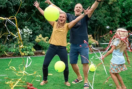 In einem Garten stehen ein Mann, eine Frau und ein Mädchen, alle in sommerlicher Kleidung. Sie werfen die Arme in die Luft und jubeln, um sie herum fliegen bunte Luftballons und Luftschlangen.