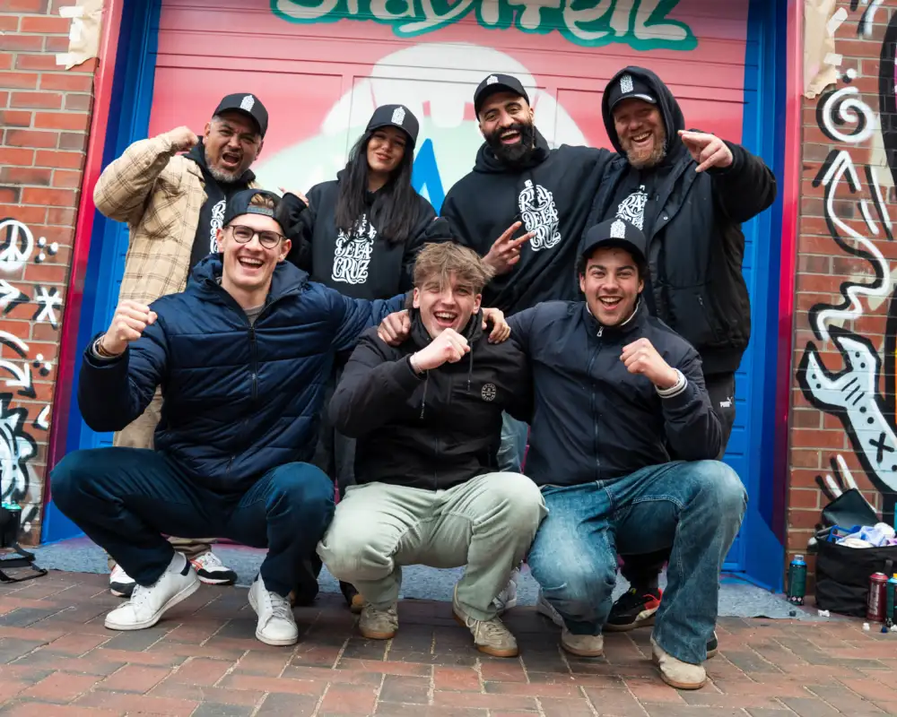 Eine Gruppe Jugendlicher steht vor einer mit bunten Graffitis besprühten Wand. Alle freuen sich und jubeln.