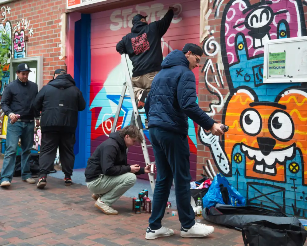 Eine Gruppe Jugendlicher steht vor einer mit bunten Graffitis besprühten Wand. Sie sind dabei, die Wand mit noch mehr Graffitis zu versehen.