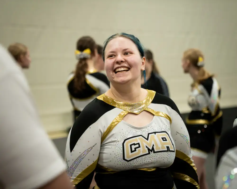 Eine jugendliche Cheerleaderin steht in ihrer Uniform in einer Sporthalle. Sie lacht jemanden an.