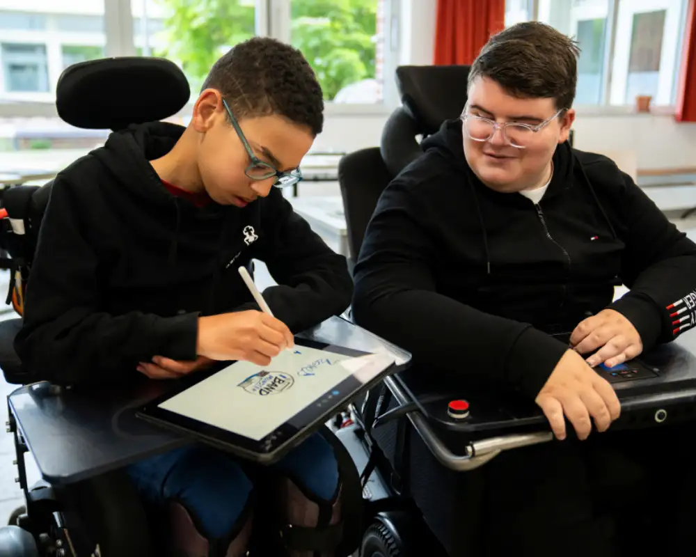 Zwei Jugendliche sitzen nebeneinander in ihren E-Rollstühlen. Einer von beiden benutzt gerade ein iPAd.