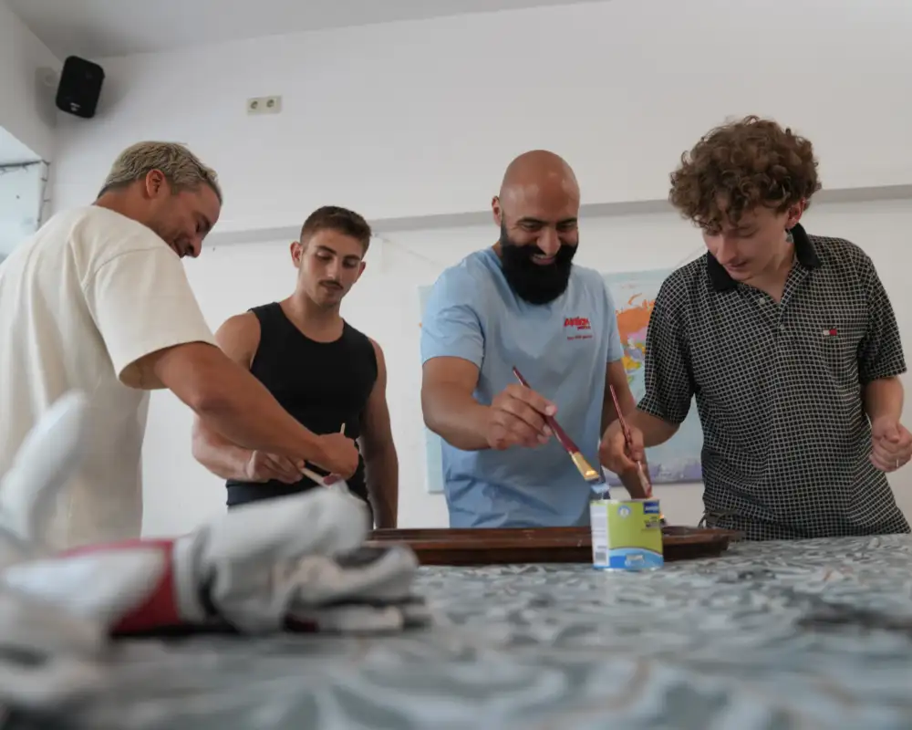 Vier junge Männer stehen an einem Tisch und bestreichen ein Möbelstück mit Farbe.