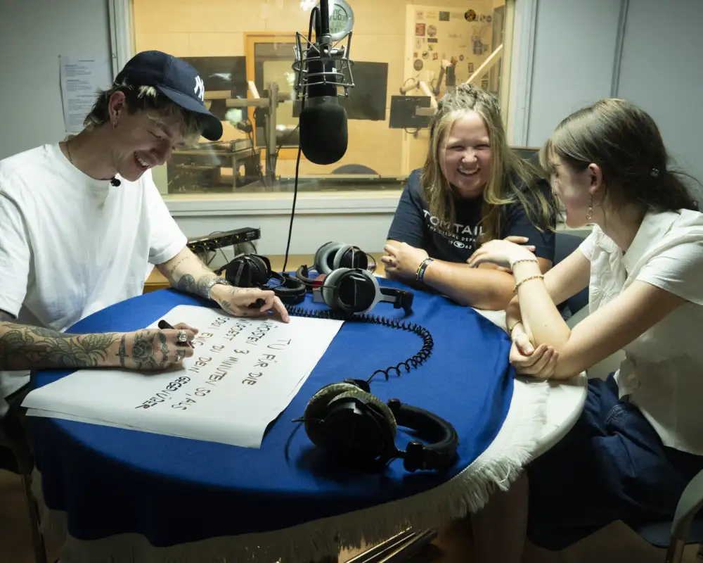 Drei junge Personen sitzen in einem Radio-Aufnahmestudio.