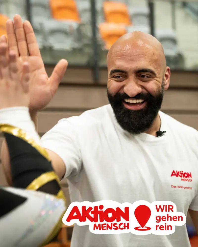 Rashid Hamid steht in einer Sporthalle und schlägt mit einer Person ein. Unten rechts sieht man das Logo der Aktion Mensch gemeinsam mit einem Location-Pin und dem Schriftzug "WIR gehen rein".