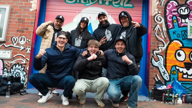 Eine Gruppe Jugendlicher steht vor einer mit bunten Graffitis besprühten Wand. Alle freuen sich und jubeln.
