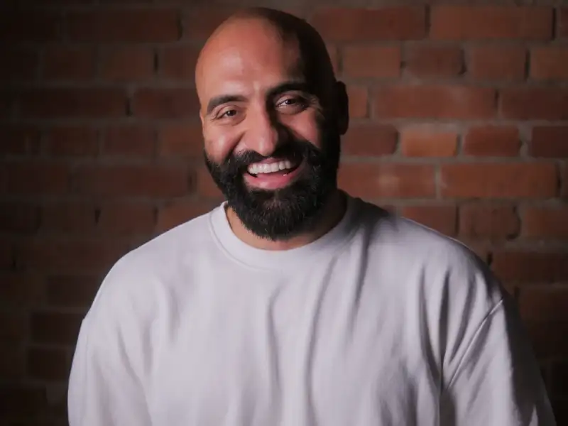 Ein Foto von Rashid Hamid. Er trägt ein weißes T-Shirt, einen schwarzen Vollbart und lacht freundlich in die Kamera.