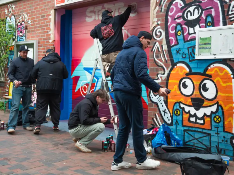 Eine Gruppe Jugendlicher steht vor einer mit bunten Graffitis besprühten Wand. Sie sind dabei, die Wand mit noch mehr Graffitis zu versehen.