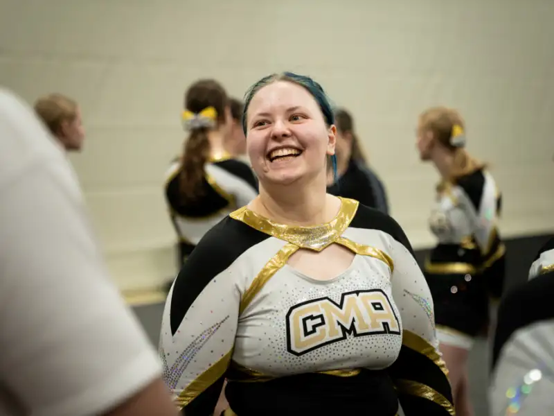 Eine jugendliche Cheerleaderin steht in ihrer Uniform in einer Sporthalle. Sie lacht jemanden an.