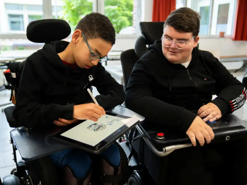 Zwei Jugendliche sitzen nebeneinander in ihren E-Rollstühlen. Einer von beiden benutzt gerade ein iPAd.