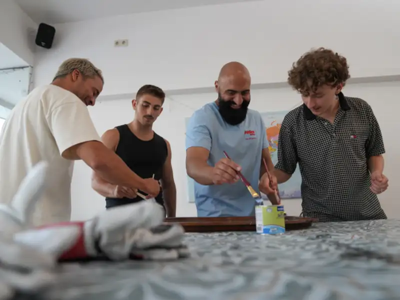 Vier junge Männer stehen an einem Tisch und bestreichen ein Möbelstück mit Farbe.
