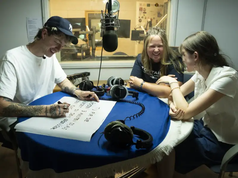 Drei junge Personen sitzen in einem Radio-Aufnahmestudio.