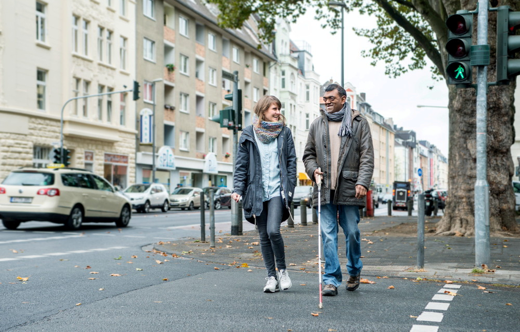 Zwei Personen gehen überqueren die Straße an einer Ampel. Der Mann führt einen Blindenstock.