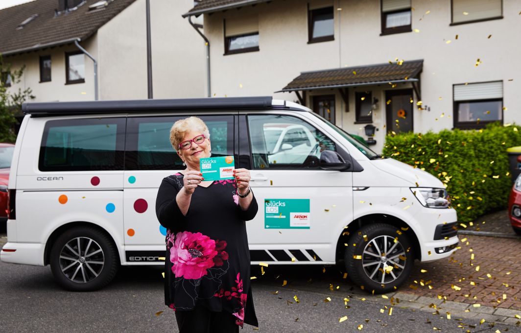 Gaby, die Gewinnerin, steht lachend vor dem Auto und hält ein Glück-Los in die Kamera.